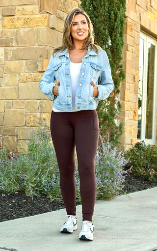 Woman wearing a light blue denim jacket, white top, and brown leggings standing outdoors.