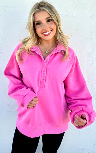 Smiling woman wearing a bright pink snap-button pullover sweatshirt with long sleeves and black leggings standing against a white textured wall.