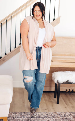 Plus-size model wearing cream sleeveless cardigan with pockets, paired with distressed jeans and boots.