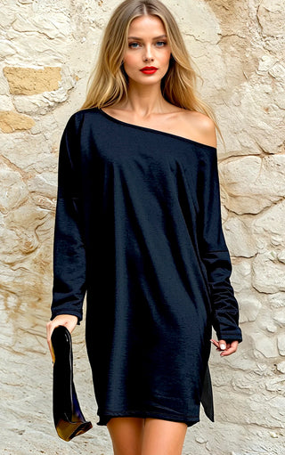 A woman wearing a black knee-length dress with a boat neckline and dolman sleeves, in an outdoor setting with a stone wall in the background.
