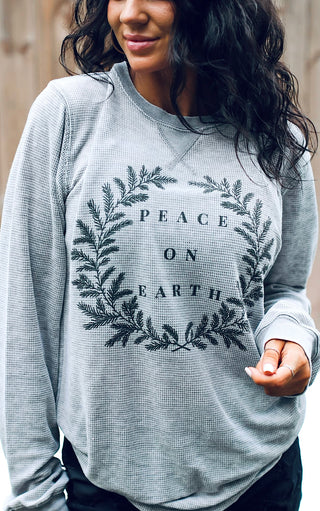 A woman wearing a grey waffle knit long-sleeve top with 'PEACE ON EARTH' screen printed in a floral design.