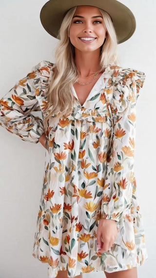 Video of a Blonde woman in a rust-and-gold floral long sleeve dress with ruffle shoulders, smiling and wearing a felt hat.
