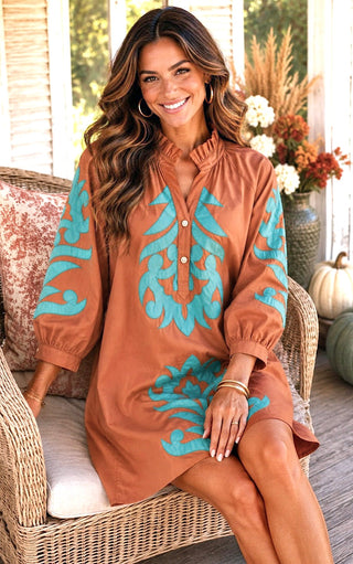 A woman sits on a wicker chair wearing a rust brown  tunic dress with turquoise blue embroidered patterns on the sleeves and chest. The setting is a warm, decorated porch with pumpkins and floral arrangements in the background.