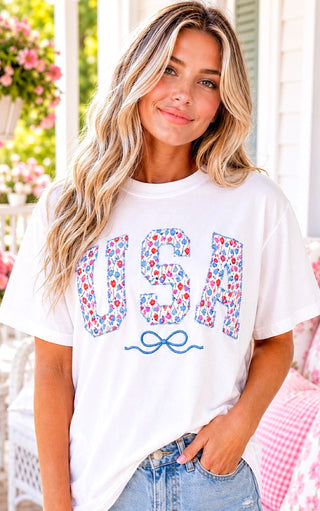 Smiling blonde model wearing a white USA t-shirt with floral tulip patch letters and an embroidered blue bow, styled with distressed denim shorts on a Southern style front porch.