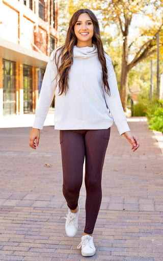 Woman walking outdoors wearing a white sweater, brown leggings, and white sneakers.