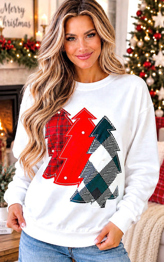 A woman wearing a white crewneck sweatshirt with a graphic of three Christmas trees in red, green, and black-and-white herringbone patterns, posed in a cozy holiday living room setting.