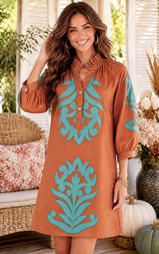 A woman wearing a rust-orange tunic dress with turquoise blue ornamental embroidery, standing in a decorated porch setting with pumpkins and wicker furniture.