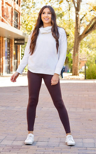 Woman wearing a white sweater and dark leggings standing on a sidewalk with trees and buildings in the background.