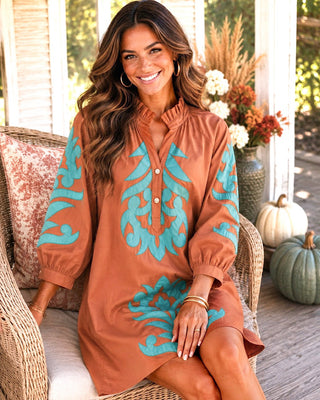 A woman wearing a rust-orange long-sleeve tunic dress with turquoise blue scrollwork embroidery, sitting on a wicker porch chair.