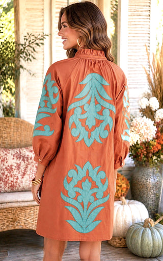 A woman wearing a rust brown long-sleeve tunic dress with large turquoise damask-style motifs on the back, photographed in a warm, decorated indoor porch setting with pumpkins and floral arrangements.