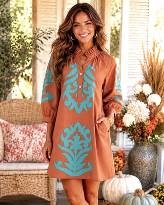 A woman modeling a rust brown  tunic dress with turquoise blue botanical patterns, standing on a decorated porch with pumpkins and floral arrangements in the background.