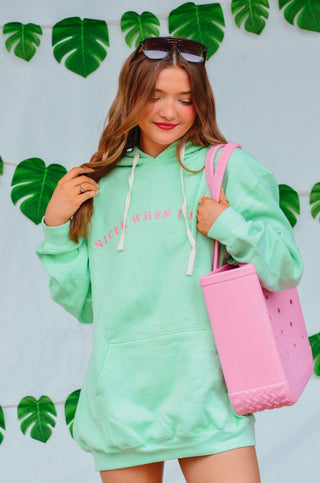 Woman wearing a light green hoodie with text, holding a pink bag against a leafy background