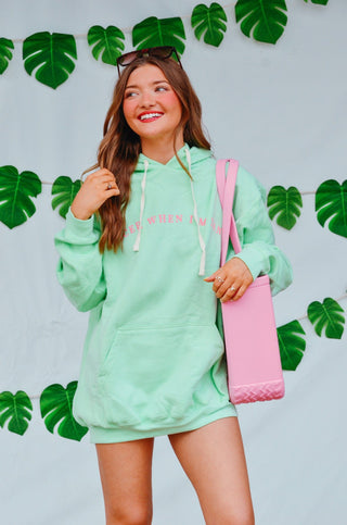 Woman wearing a light green hoodie and shorts set with a pink bag, standing against a light gray background with leaf decorations.