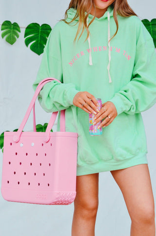 Person wearing a light green hoodie with text, holding a pink perforated tote bag and colorful drink.