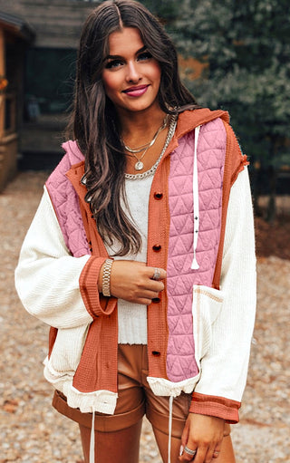 Woman wearing a pink and orange quilted vest over a white sweater and brown pants.