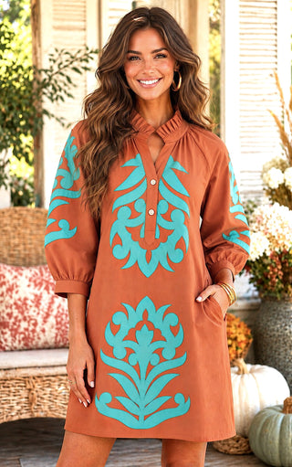 A woman modeling a toffee brown shift dress with turquoise damask-style patterns on the sleeves and front, set against a warm, autumnal porch background with pumpkins and wicker.