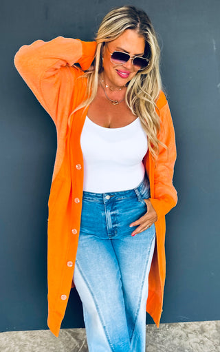 Woman wearing an orange cardigan, white top, and blue jeans against a gray wall.