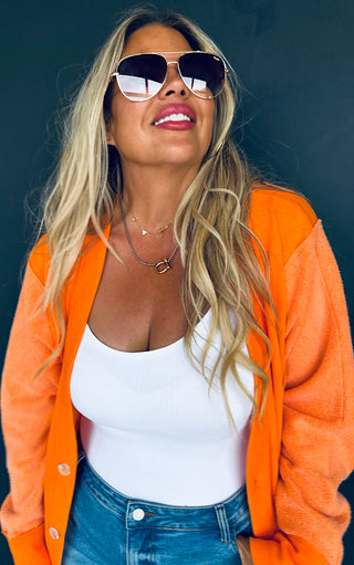 Woman wearing an orange cardigan, white top, and blue jeans with sunglasses against a dark background