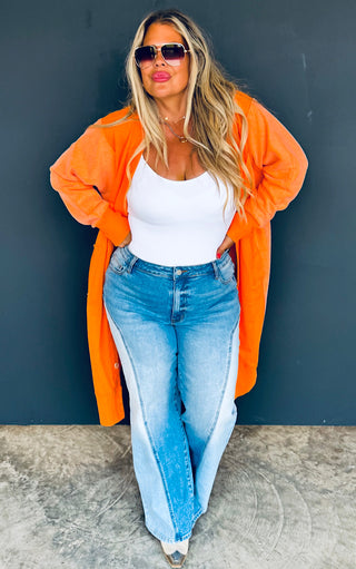 Woman wearing an orange cardigan, white top, and blue jeans against a dark background