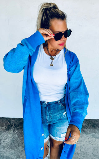 Woman wearing a blue cardigan, white top, and denim shorts against a plain background