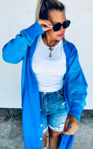 Woman wearing a blue cardigan, white shirt, and denim shorts against a plain background