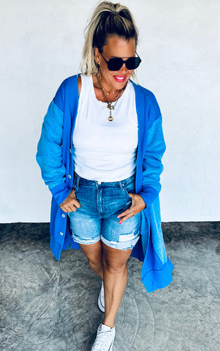 Woman wearing a blue cardigan, white top, denim shorts, and sunglasses against a white wall.
