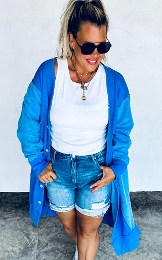 Woman wearing a blue cardigan, white top, and denim shorts against a plain background