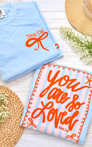 Light blue t-shirt with 'John 3:16' and ‘You are so loved’ text lying on a wooden background with spring accessories 