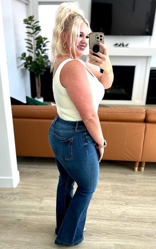 Blonde woman wearing dark wash Judy Blue trouser flare jeans and a white tank top taking a selfie to show fit from the side
