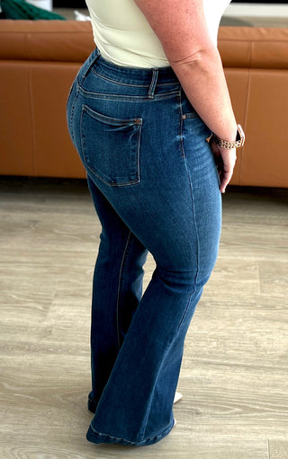 Side view of woman wearing blue flare jeans standing on a wooden floor with a brown leather couch in the background.