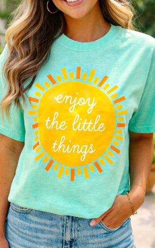 Person wearing a mint green t-shirt with a yellow sun graphic and text 'enjoy the little things'.