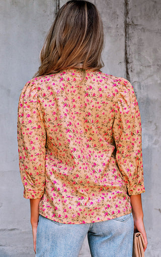 Back view of Woman wearing a golden floral blouse with pink flowers, pintuck pleats, three bow tie neckline, and three-quarter puff sleeves, standing against a light grey textured wall with one hand on her hip.