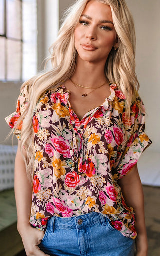 Blonde woman wearing the Fields of Color Top, a short-sleeve floral blouse with ruffle neckline and tie detail, paired with blue jeans.