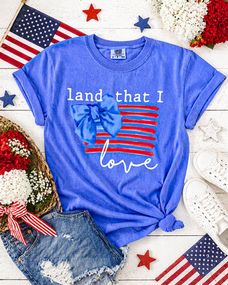 Blue t-shirt with 'land that I love' design on a wooden surface with American flags and flowers.