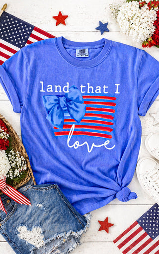 Blue t-shirt with 'land that I love' design on a wooden surface with American flags and flowers.