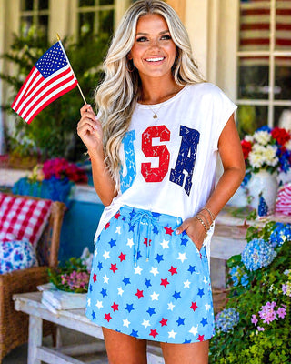 Woman holding an American flag, wearing a white t-shirt with 'USA' and a blue skirt with red and white stars, outdoors.