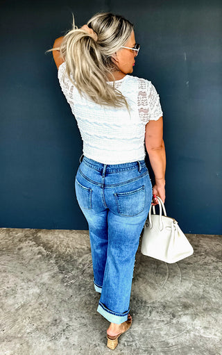 Woman in white lace top and blue jeans holding a white handbag against a dark blue wall.