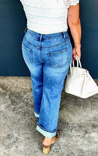 Person wearing blue jeans and a white top, holding a white handbag against a dark blue wall.