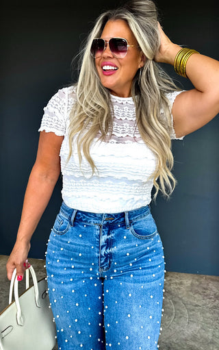 Woman wearing a white lace top and pearl studded blue jeans with sunglasses and a handbag.