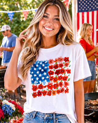 Woman wearing a white t-shirt with a colorful American flag design, standing outdoors with an American flag in the background.