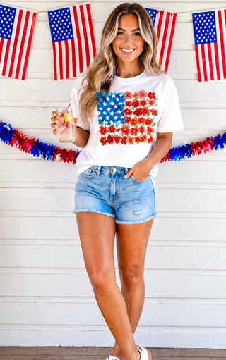 Woman wearing a white t-shirt with an American flag design and denim shorts, standing in front of American flags and bunting.