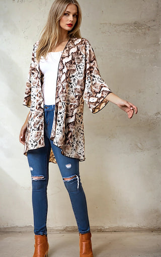 Woman wearing a patterned kimono, white top, blue jeans, and brown boots against a plain wall.