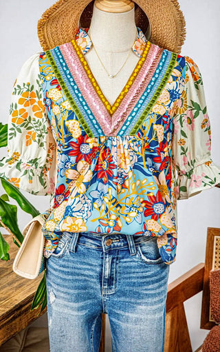 Mannequin wearing a blue floral V-neck blouse with colorful trim and puff sleeves, styled with denim jeans.