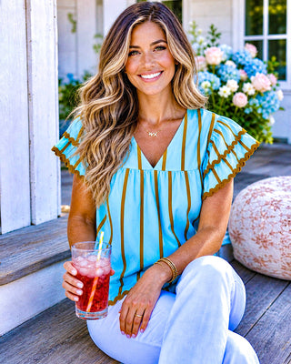 model wearing robin’s egg blue blouse with brown stripes and rickrack trim with white jeans holding a drink on a Southern porch 