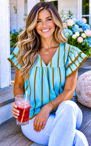 Seated  view of model wearing robin’s egg blue blouse with brown stripes and rickrack trim with white jeans