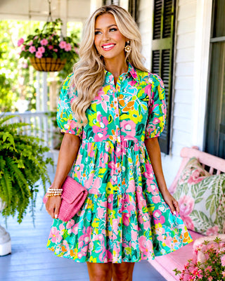  Blonde model in a vibrant pink floral tiered dress standing outdoors beside pink flowers and porch seating