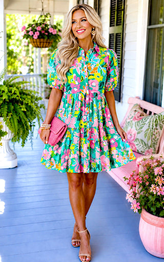 Southern blonde model posing in a bright floral dress with pink clutch and cottage porch background.