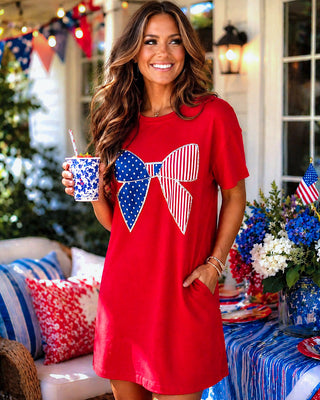 Woman wearing a red dress with a large bow design, holding a drink, in a festive outdoor setting.