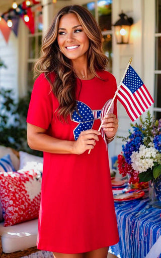 Woman in a red dress holding small American flags in a decorated outdoor setting with flowers and flags.