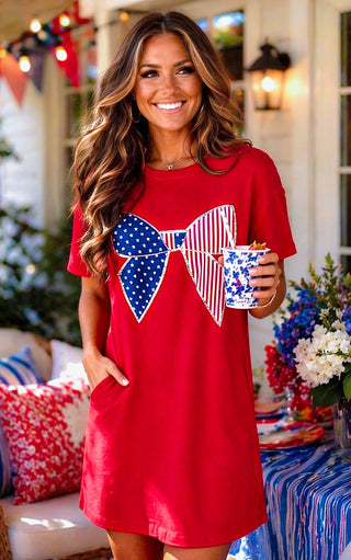Woman wearing a red dress with an American flag bow design, holding a mug, in a festive outdoor setting.